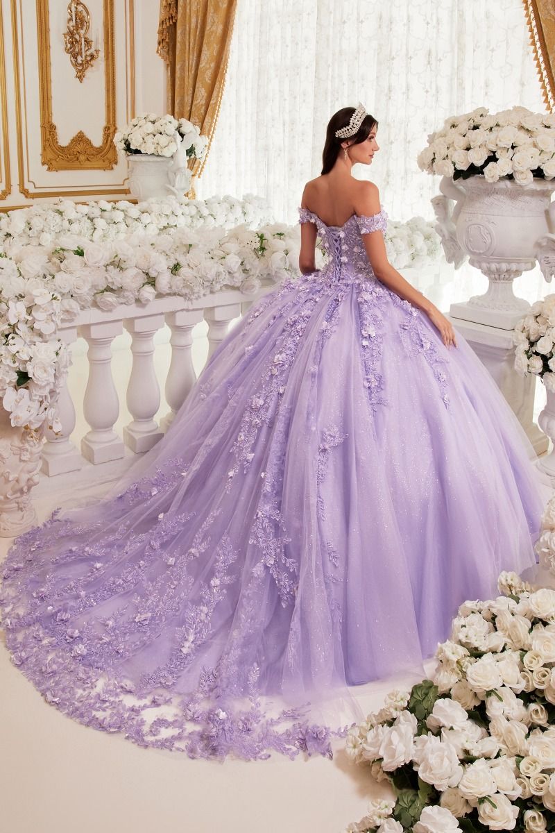 A woman in the Off-Shoulder Purple Quinceanera Dress: Ladivine 15717 stands facing away in a flower-filled room, her shimmering a-line ball gown with floral details glowing under the light.